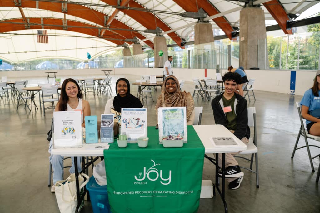 Four Joy Project Volunteers sitting behind a table for The Joy Project for the NEDA Care Fair. The table is covered with a green cloth with The Joy Project branding. The table displays flyers, brochures, and other giveaway items.